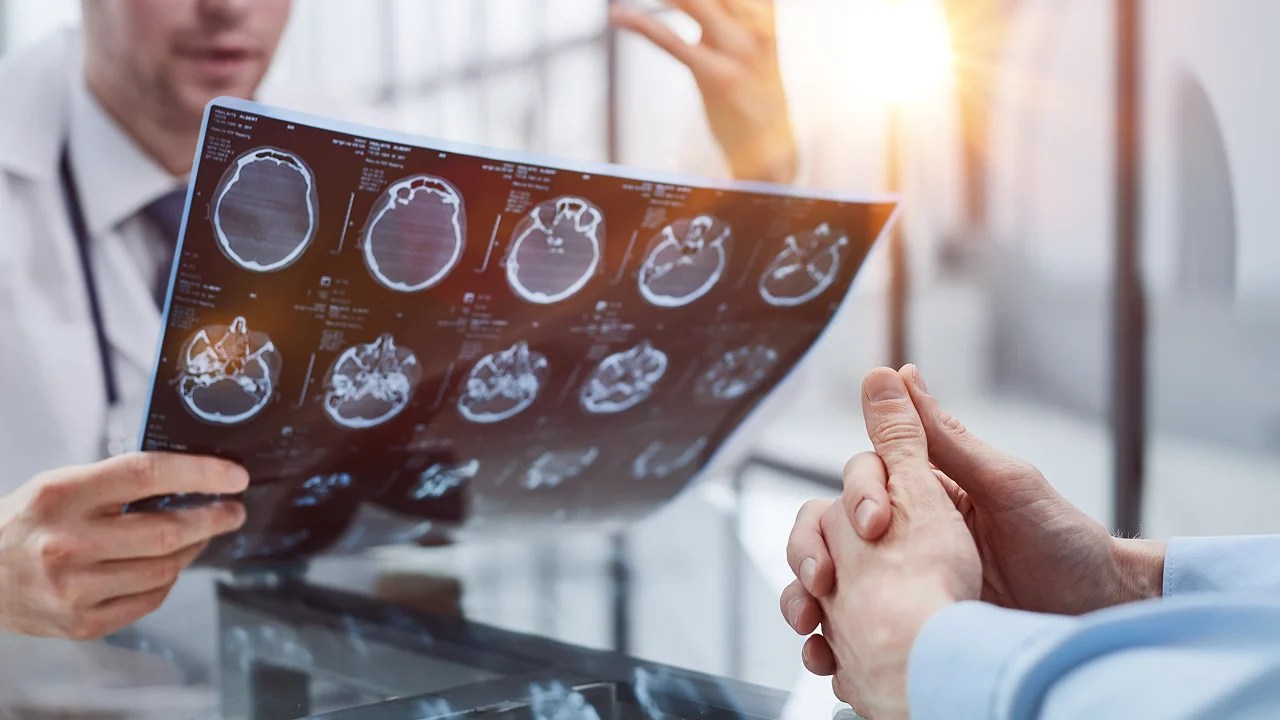 A doctor holding up and examining a brain scan film with a patient seated across from them.