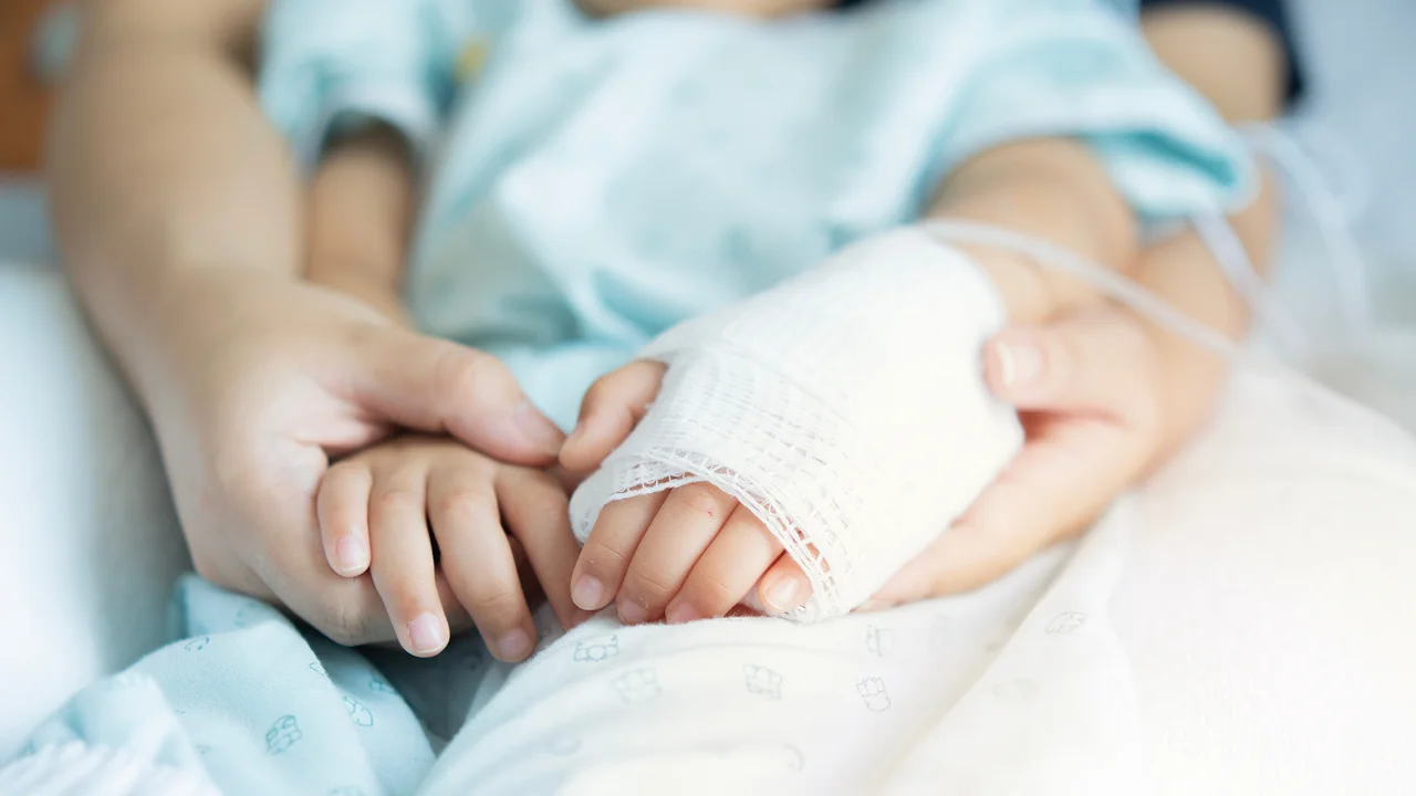Adult holding a child's bandaged hand with IV line in a hospital setting.