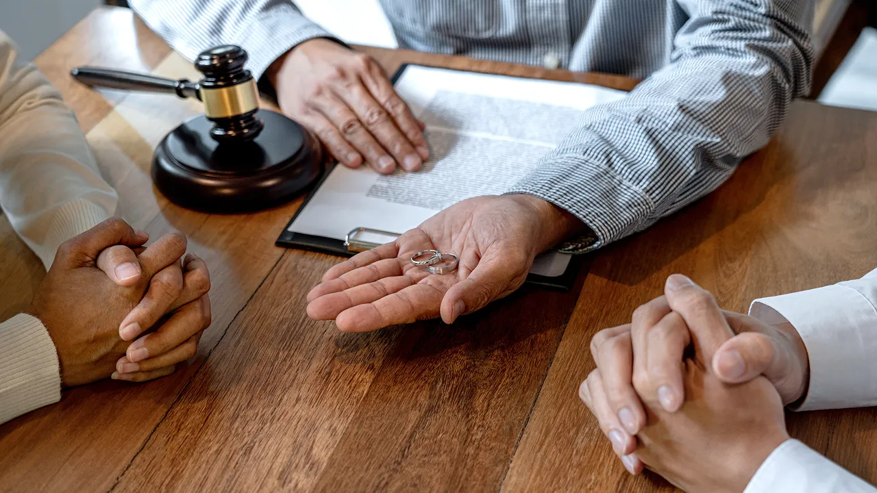 An open hand holding two wedding rings over a wooden table, with a gavel and clipboard in the background.