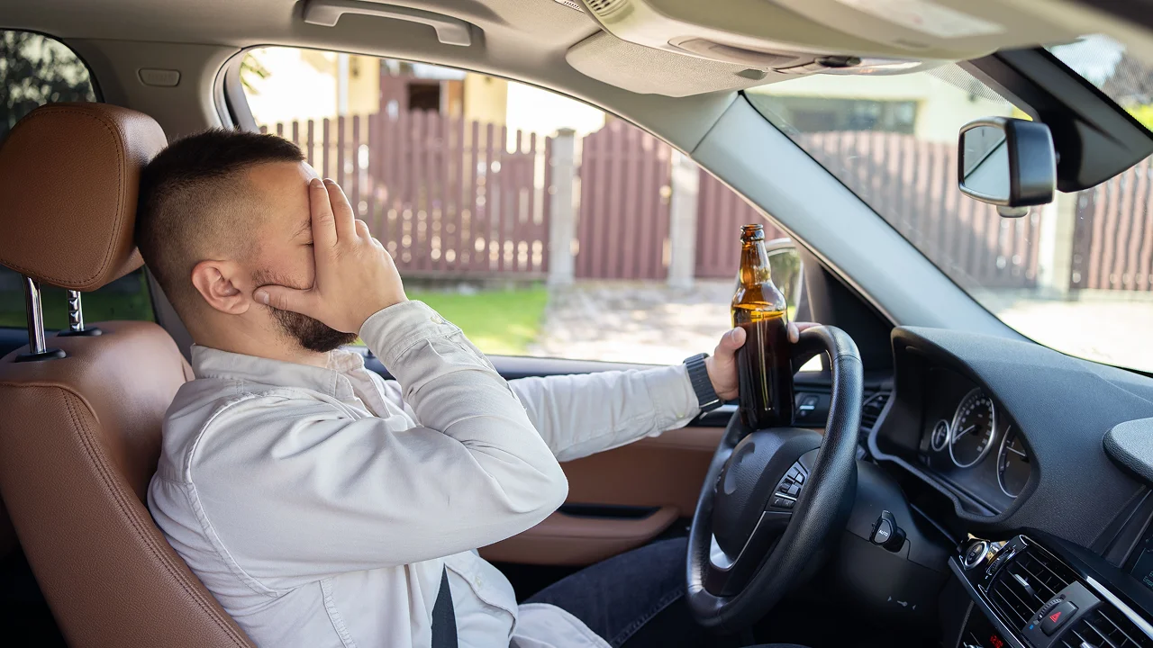 Drunk driver covering his face while holding a beer bottle at the wheel.