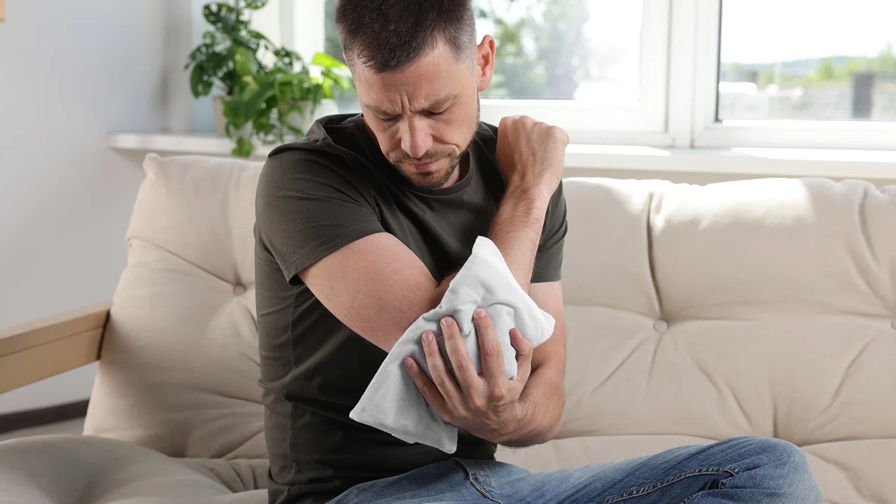 A man sitting on a sofa applying a cold compress to his elbow.