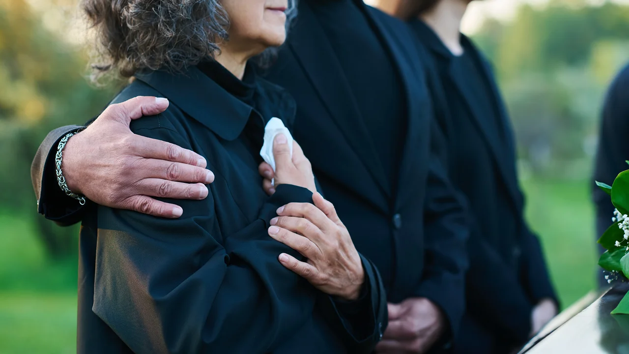 A comforting hand on a grieving woman's shoulder at an outdoor funeral.
