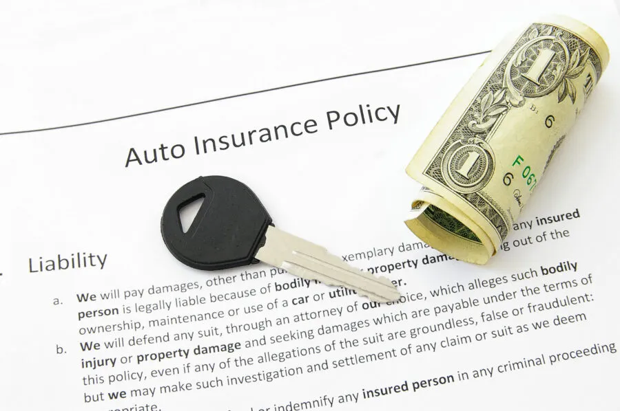A car key and a roll of US dollar bills resting on an auto insurance policy document.