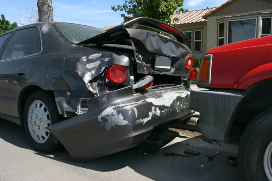 A red vehicle rear-ending a dark grey sedan, causing significant damage to the sedan's trunk and bumper.
