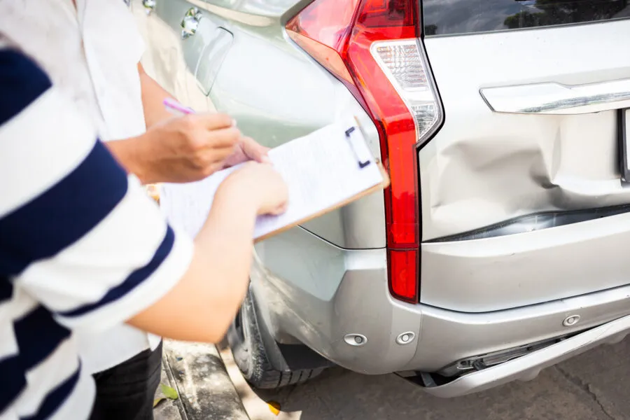 An insurance agent writing on a clipboard while inspecting a large dent in the rear bumper of a silver SUV.