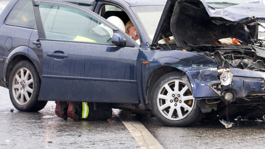 Crash. A car with a broken hood on the road. An injured passenger is sitting in the cab. She is being assisted by a lifeguard.