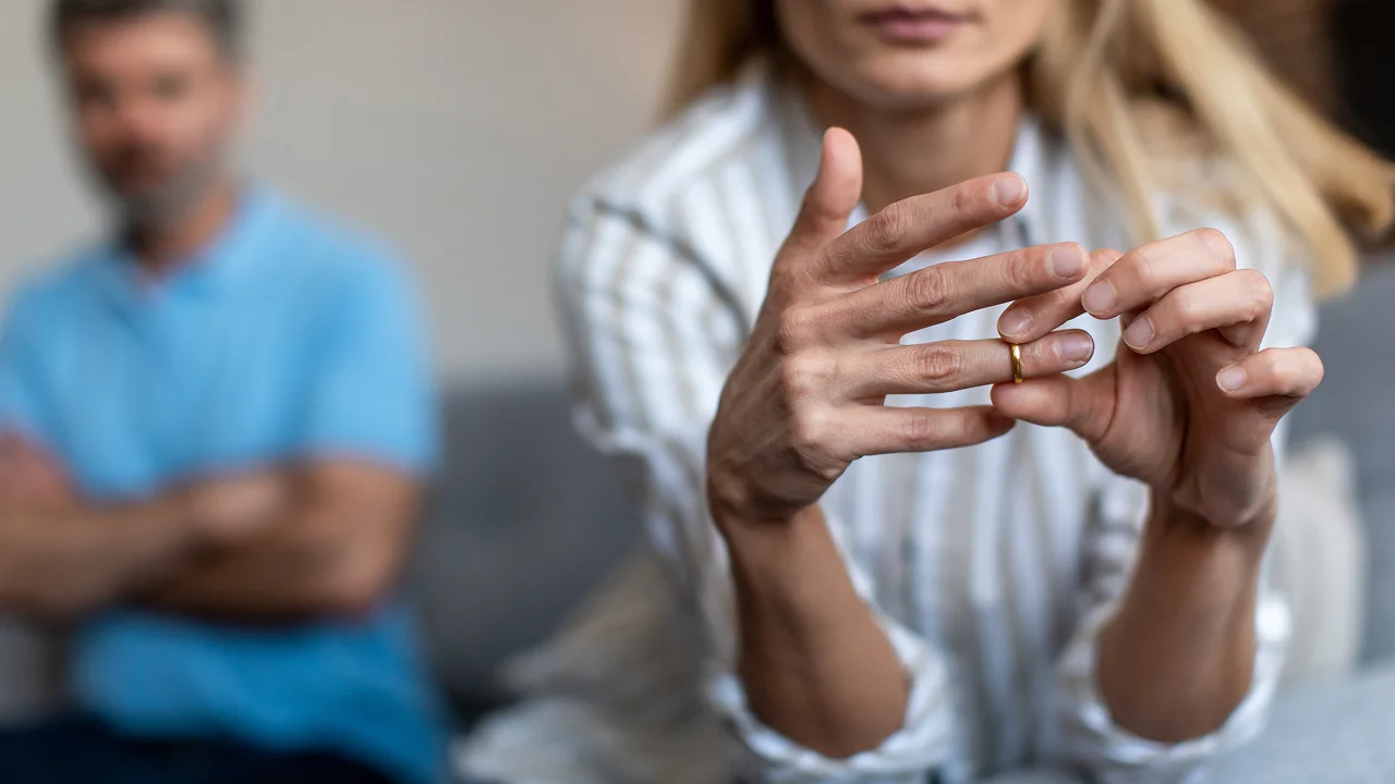 Woman taking off her wedding ring with a blurred man sitting in the background.