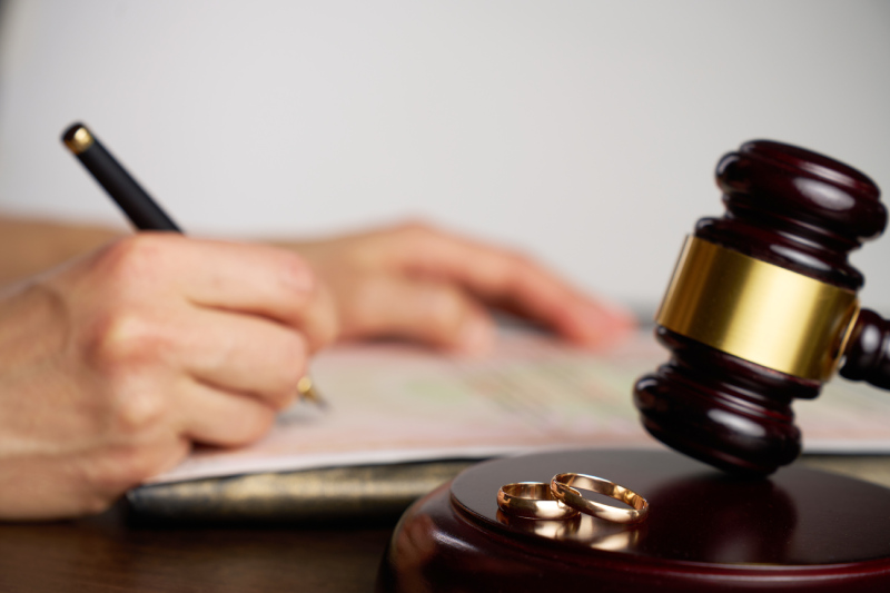 Close-up of a person signing divorce document, with a judge gavel and wedding rings in focus. A symbolic representing the legal process of divorce, separation agreements, or prenuptial documentation