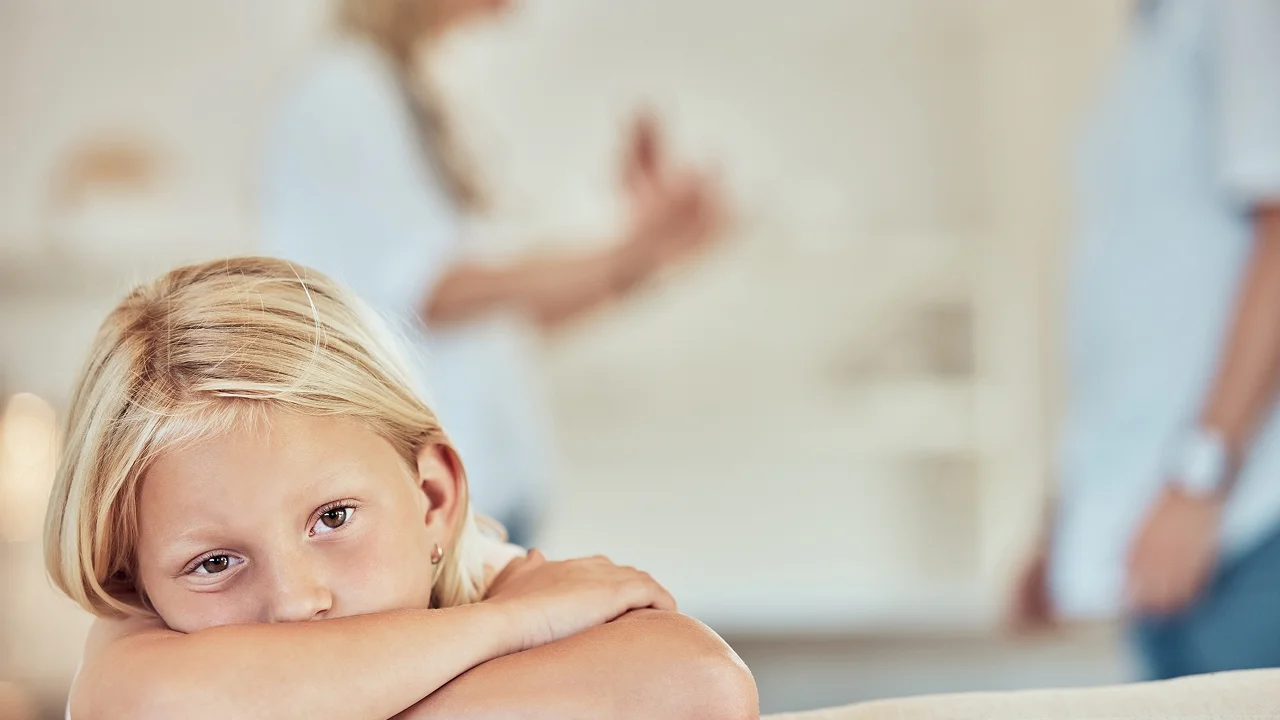 Sad girl resting her head, with arguing adults blurred in the background.